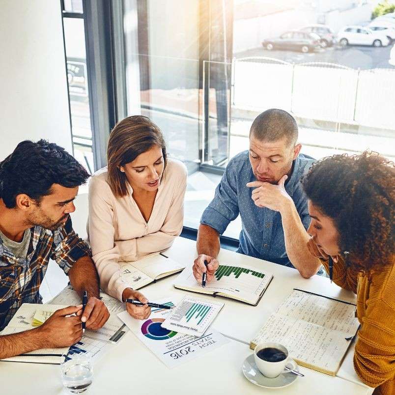 Four people sit around a table in a bright office, discussing documents, charts, and an Agile Project Charter Template. With notebooks and coffee in hand, they appear focused and engaged in a collaborative meeting.