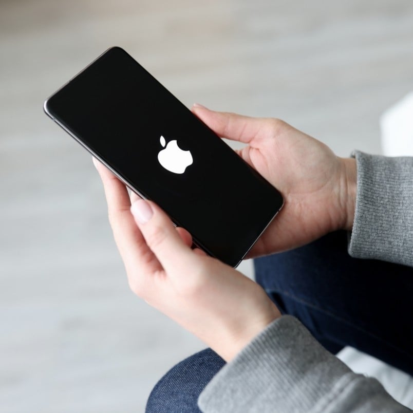 A person holding a black iPhone with the Apple logo displayed on the screen, suggesting the phone is starting up—an agile project management example of staying connected. The person is wearing a gray sleeve and blue jeans.