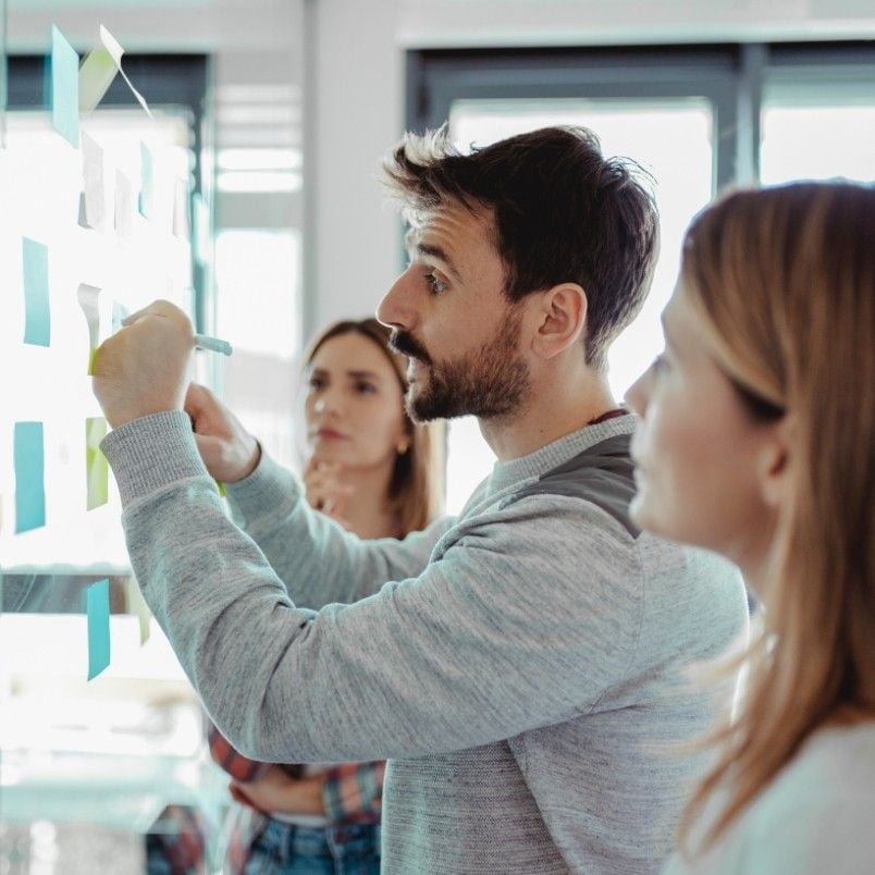 Three people stand by a glass wall covered in sticky notes. A man writes on a note while two women look on, demonstrating agile skills as they engage in a focused collaborative discussion.