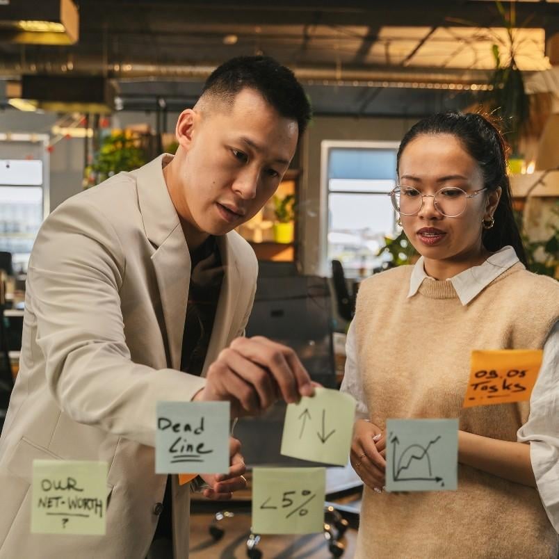 Two colleagues in an office discuss notes and charts on glass. One, highlighting the project manager role in scrum, points at sticky notes with phrases like "Deadline," graphs, and "<5%," while the other listens attentively. Plants and desks are visible in the background.
