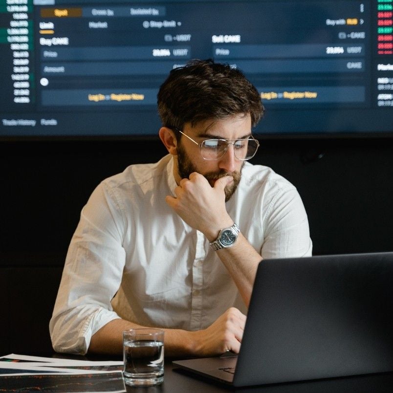 A man with glasses and a beard sits at a desk, looking thoughtfully at a laptop. Nearby, documents and a large screen displaying stock market data suggest he’s engaged in alternative analysis.