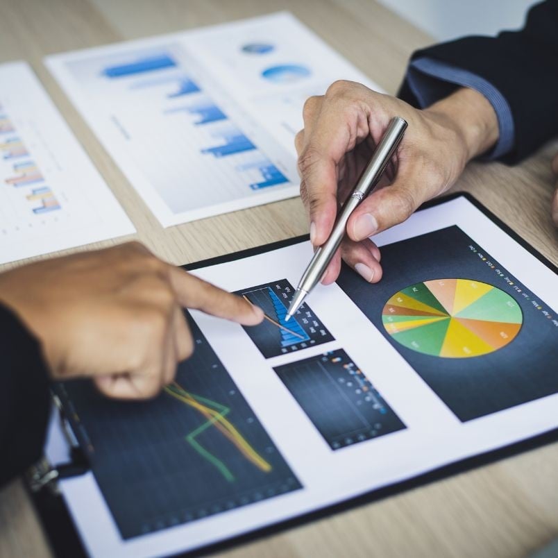 Two people discuss financial charts and graphs on printed documents. One person holds a pen, pointing to a bar chart, while the other references a risk register alongside data on a clipboard.