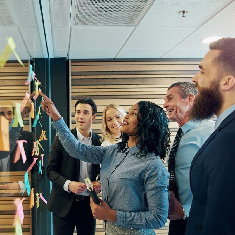 A group of four diverse professionals smile and discuss ideas while standing by a glass wall covered with colorful sticky notes, collaborating on a project communication plan in a modern office setting.
