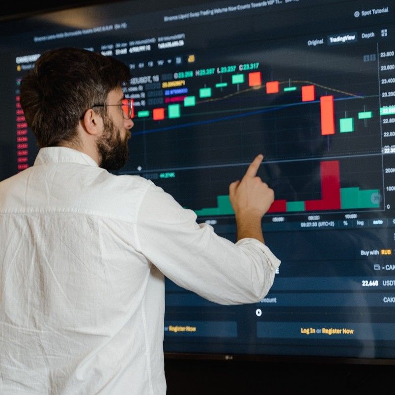 A man in a white shirt points at a large screen displaying a colorful candlestick stock chart, trading information, and project management workflow details.