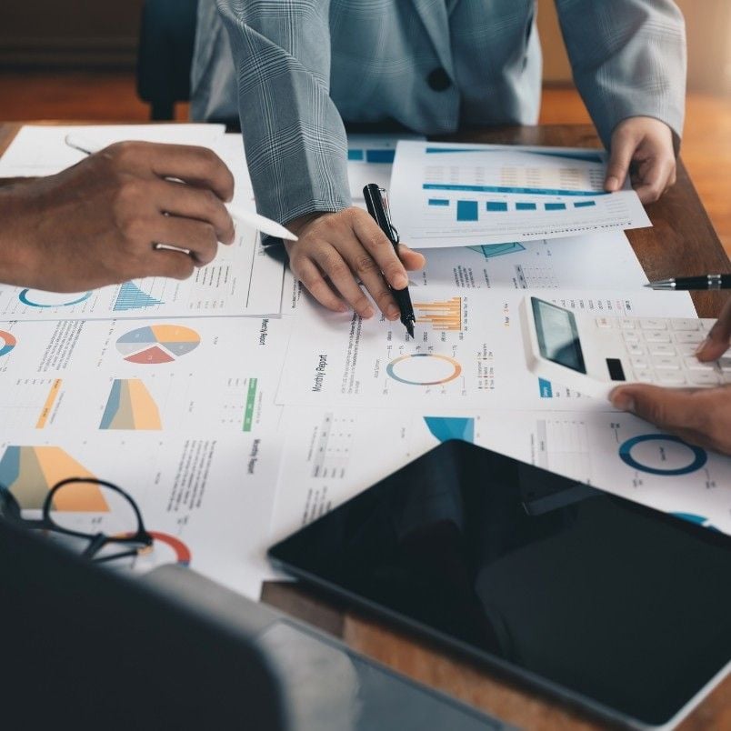 Three people analyze and discuss financial charts and graphs spread out on a table, using pens and a calculator, with a digital tablet nearby, as they streamline project management business process strategies.
