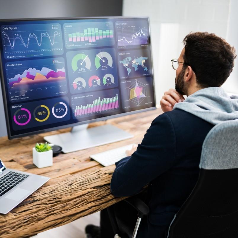 A person sits at a desk in a modern office, looking at a large computer monitor displaying colorful charts, graphs, and data visualizations as part of their project management workflow.