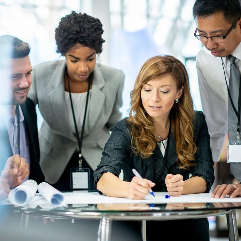 Four business professionals gather around a table, collaborating and reviewing documents. A woman is seated, writing on paper, while three colleagues stand closely, discussing project management strategies and streamlining the business process.