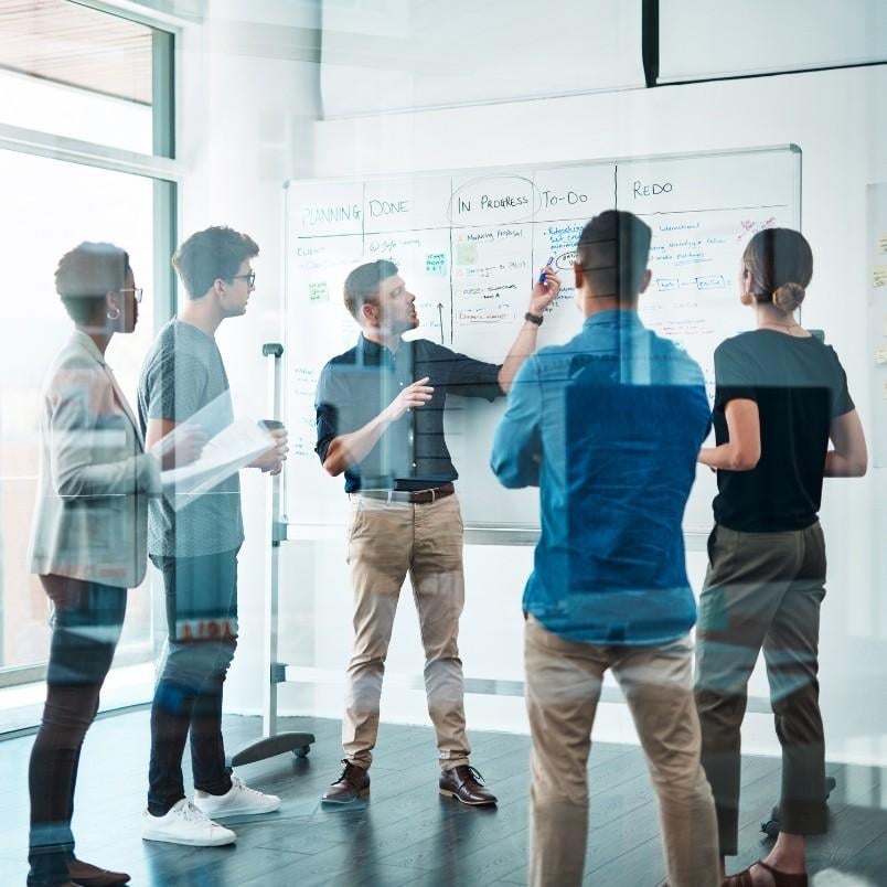 Five people stand around a whiteboard covered with notes and diagrams, discussing the project manager role in Scrum and brainstorming in a modern office space with glass walls.