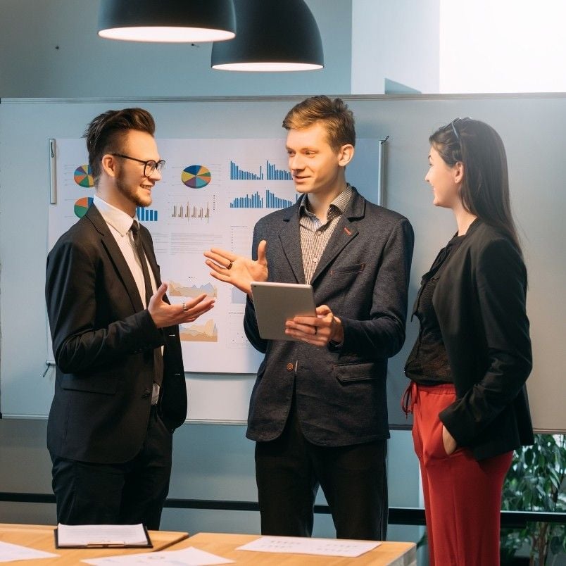 Three young professionals in business attire stand and converse in front of a whiteboard with charts and graphs, illustrating an agile project management example as they discuss strategies and review data together.