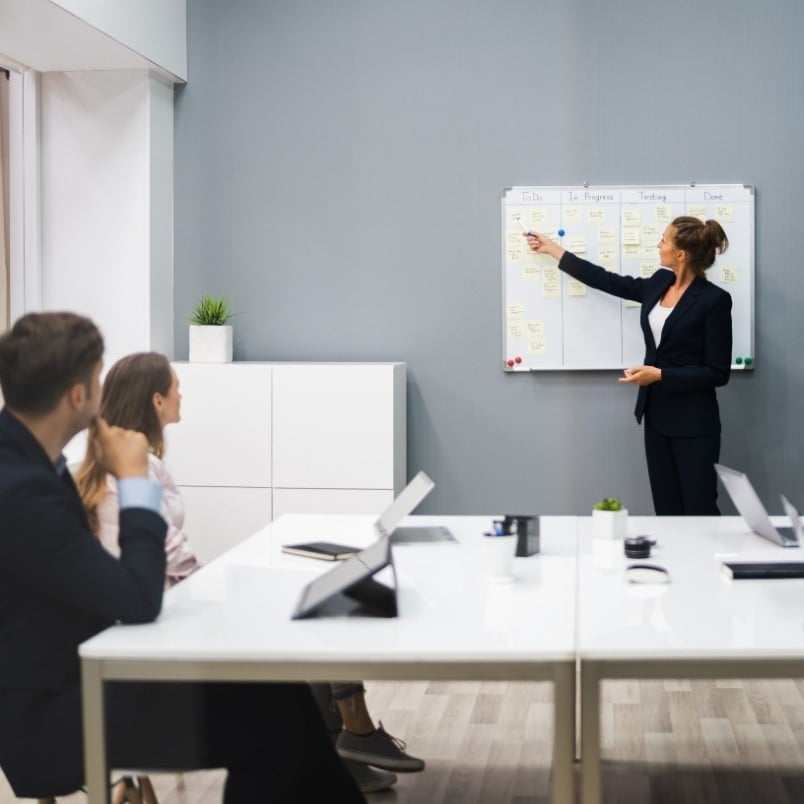 A woman in business attire stands by a whiteboard covered with sticky notes, presenting an agile project charter to three colleagues seated at a conference table in a modern office. Laptops and notebooks are on the table.