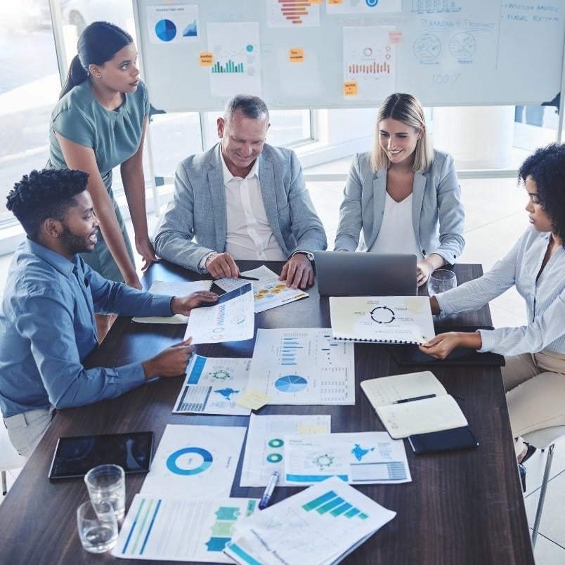 Five people sit and stand around a table covered with charts, graphs, reports, and a wireframe template, discussing work. A whiteboard with more charts is visible in the background, indicating a business meeting or brainstorming session.
