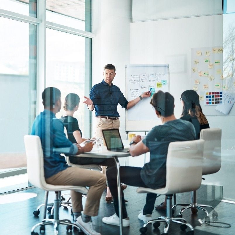 A man stands by a whiteboard presenting a story map template to four seated colleagues in a modern glass-walled meeting room, with laptops and notes on the table and sticky notes on the wall.