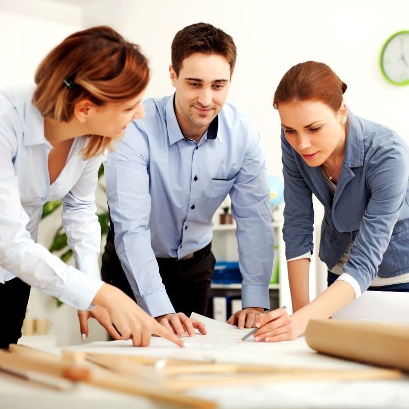Three people in business attire stand around a table, collaborating on documents and plans—including an Agile Project Charter Template—spread out before them in a bright office.
