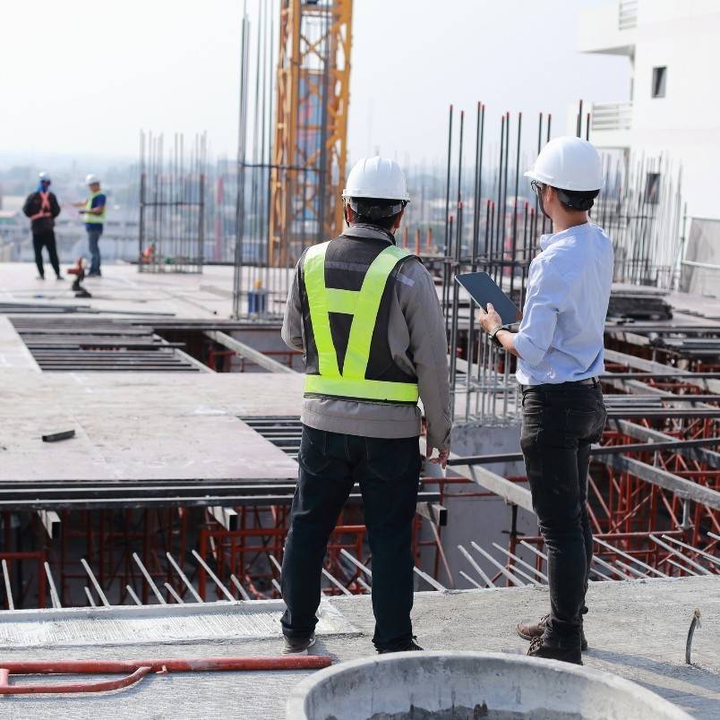 Two construction workers in safety vests and helmets stand on a building site, reviewing agile phases of project management on a tablet. Metal rods and materials surround them, with more workers visible in the background.