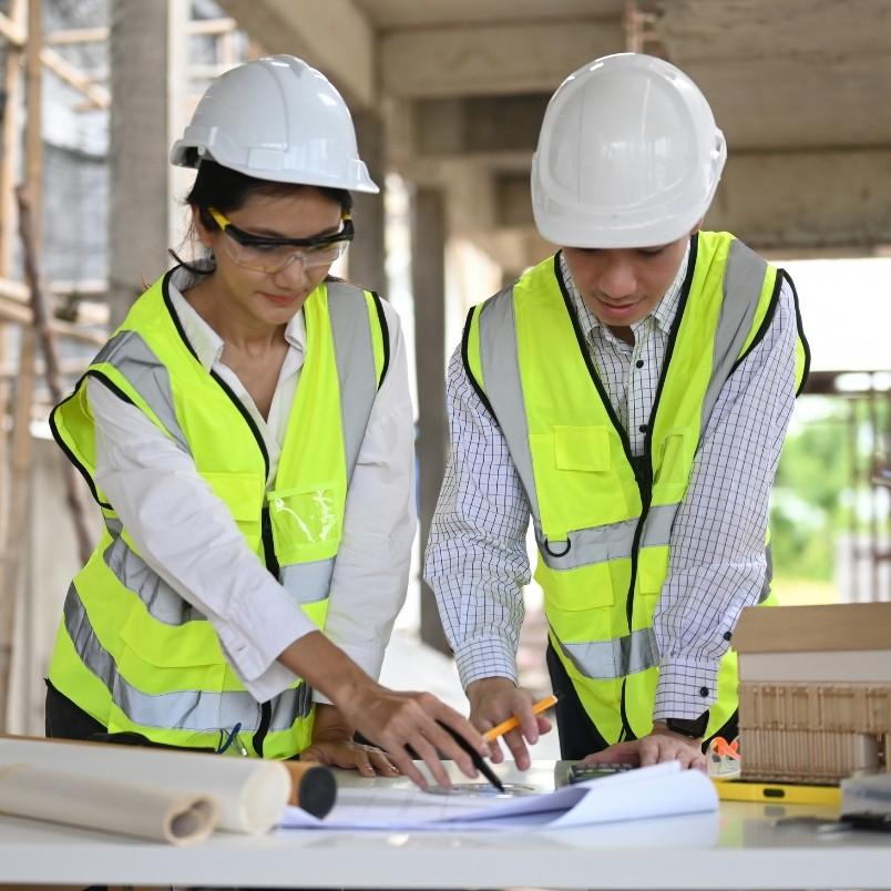 Two construction workers in white hard hats and yellow safety vests review blueprints at a construction site, discussing project details and Construction Project Management. One points at the plans while the other takes notes.