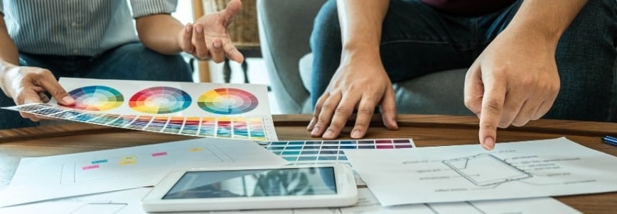 Two people sit at a table reviewing color swatches, design sketches, and an Agile Project Charter Template on a tablet, collaborating on a creative project. One gestures with a hand while the other points to a paper with a drawing.