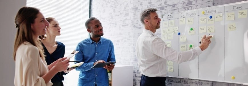 Four people in business attire stand by a whiteboard with sticky notes organized in columns, illustrating an agile project management example. One man writes on the board while the others observe, holding a tablet and notepads as sunlight streams through a window.