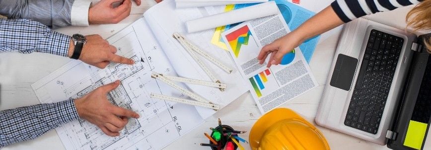 Two people at a desk review blueprints, charts, and documents. A laptop, yellow hard hat, and colorful pens suggest collaborative construction project management or architectural planning in progress.
