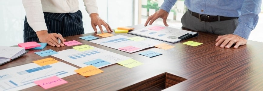 Two people stand at a wooden table covered with charts, graphs, a features template, and colorful sticky notes, appearing to discuss business data and strategies. One person is holding a folder with printed graphs.