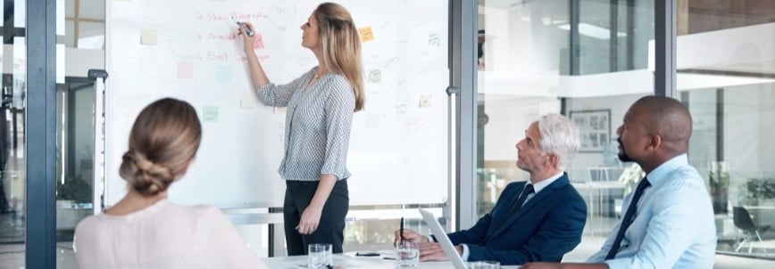A woman stands and writes on a whiteboard during a meeting, sharing a wireframe template while three colleagues seated at a table watch and listen attentively in a modern office setting.