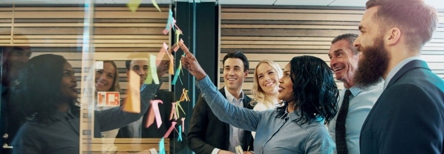 Five business professionals stand together, smiling and looking at sticky notes on a glass wall, as one woman points at a note during a collaborative brainstorming session in a modern office setting.