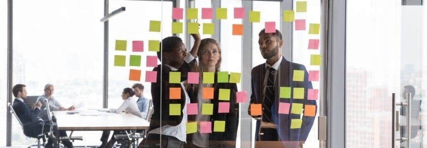 Three business professionals with agile skills look at a glass wall covered in colorful sticky notes, brainstorming in a modern office, while two other colleagues meet in the background.