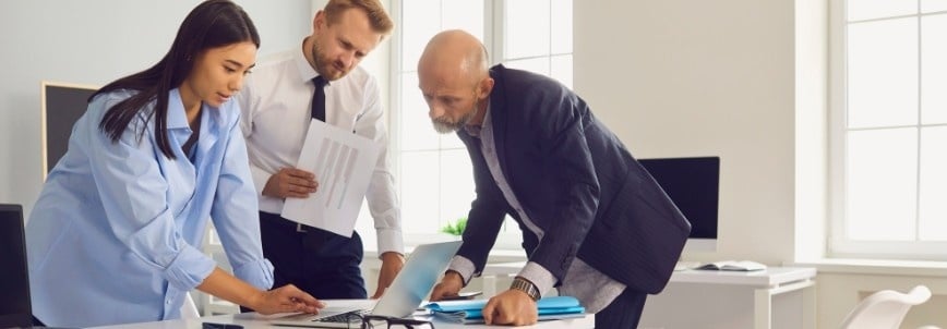 Three professionals collaborate in an office, reviewing a risk register and documents while looking at a laptop on the desk. One woman points at the laptop as two men observe, one holding papers. Sunlight streams through large windows in the background.