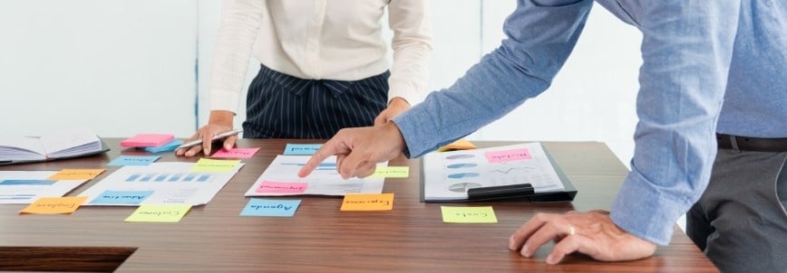 Two people stand at a table covered with papers, charts, and colorful sticky notes, discussing and pointing at documents—suggesting a collaborative planning session focused on an agile project charter.