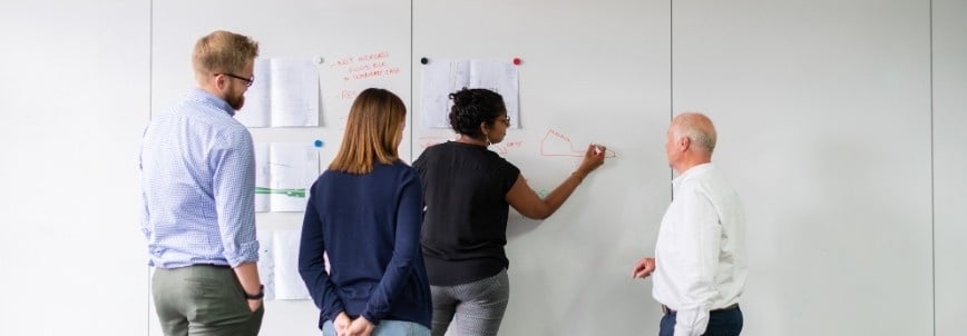Four people stand facing a whiteboard with papers and diagrams pinned to it; one person is writing on the board while the others observe and discuss an alternative analysis.
