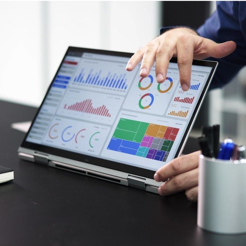 A person points at a laptop screen displaying colorful charts, graphs, and data visualizations within a wireframe template, while sitting at a desk with a pen holder and other office supplies nearby.