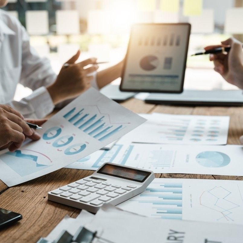 People having a business meeting, analyzing financial charts and graphs on paper and a tablet as they discuss project management and business process. A calculator and documents with data are spread on the wooden table, sunlight streaming through a window in the background.