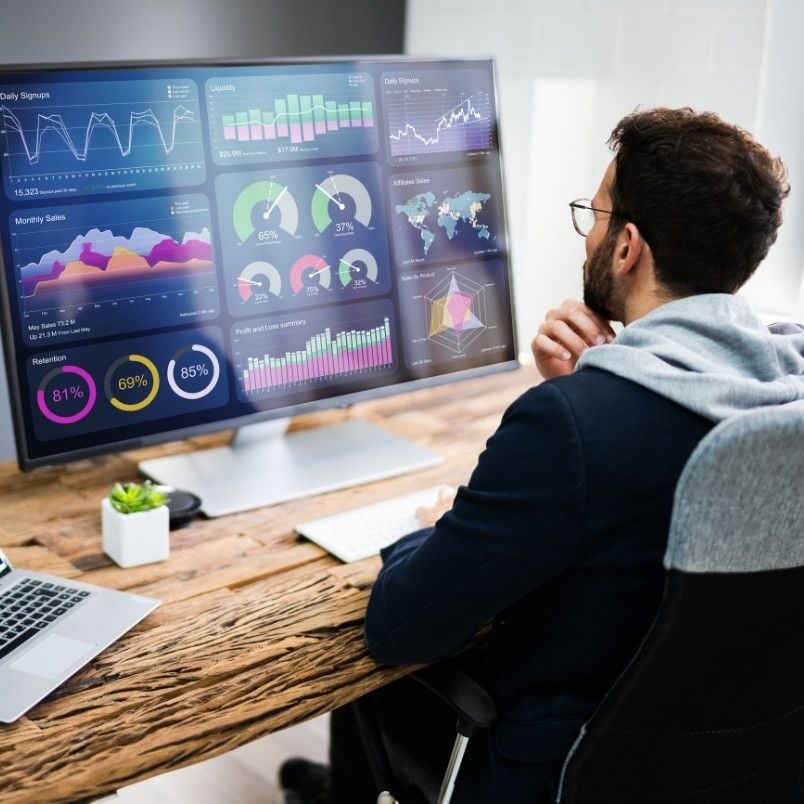 A man sits at a wooden desk, analyzing colorful data charts and graphs displayed on a large computer monitor. The screen features various analytics, including sales, growth, and a wireframe template alongside world maps.
