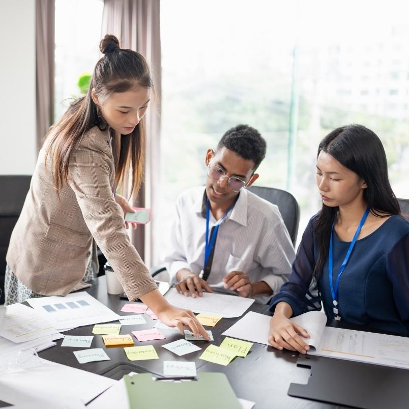 Three colleagues sit at a table covered with papers and sticky notes while one woman stands, pointing at the notes and discussing ideas with the others, illustrating a collaborative project management workflow in a bright office setting.