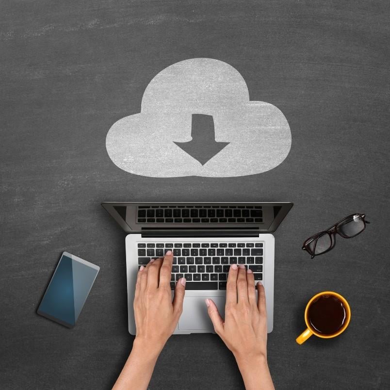 A person typing on a laptop with an Agile Team Charter concept and a cloud download icon drawn in chalk on a blackboard, next to a smartphone, eyeglasses, and a cup of coffee.