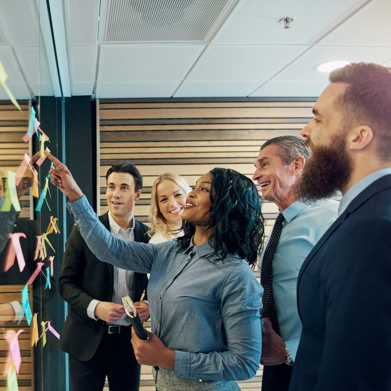 A group of five business professionals with agile skills smiling and collaborating, standing by a glass wall covered with colorful sticky notes in a modern office setting.
