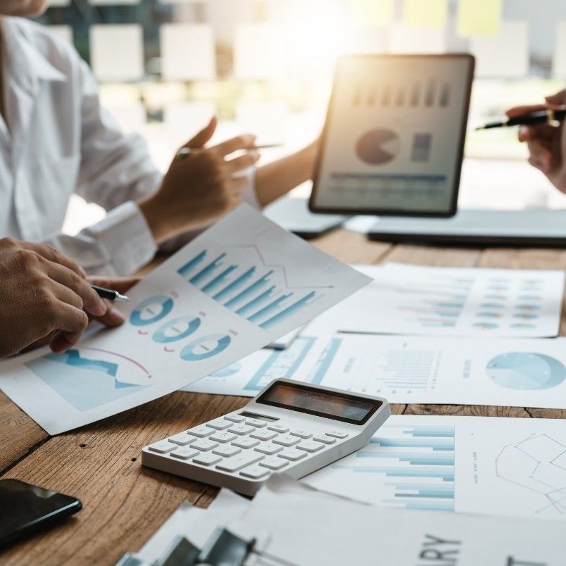 Three people discuss graphs and charts at a table covered with financial documents, a calculator, and a tablet displaying charts, collaborating on project management workflow as sunlight shines in the background.
