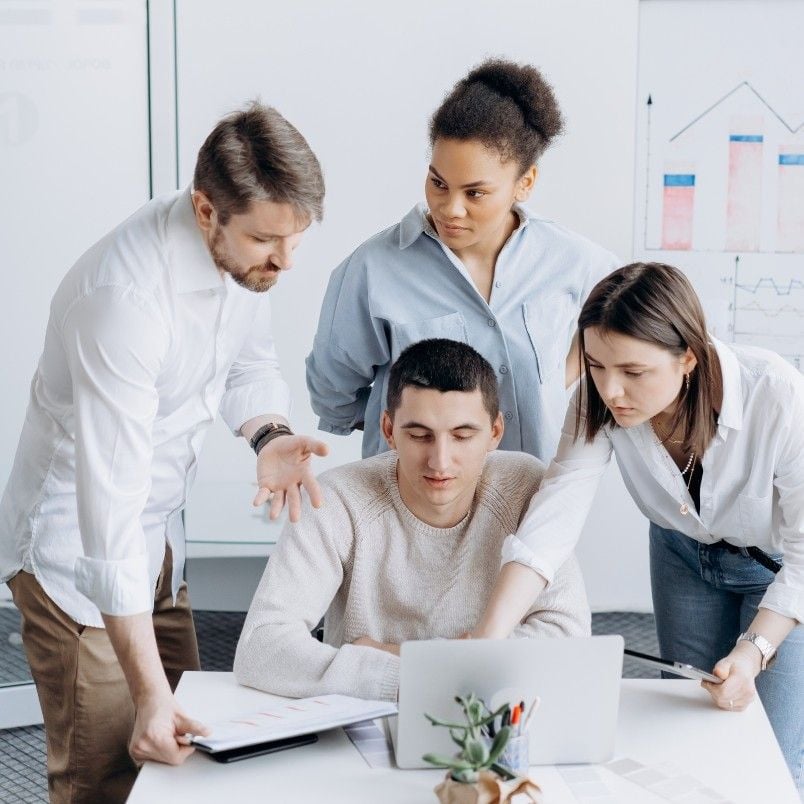 Four people gather around a laptop in an office, collaborating on a project communication plan. Three stand and one sits, all focused on the screen. A document and charts are visible in the background.
