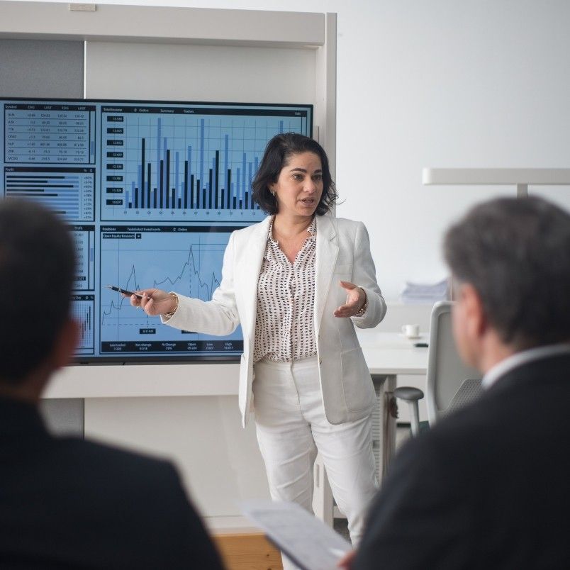 A woman in a white suit presents root cause analysis data on large screens displaying graphs and charts to a group of people in a modern office setting.