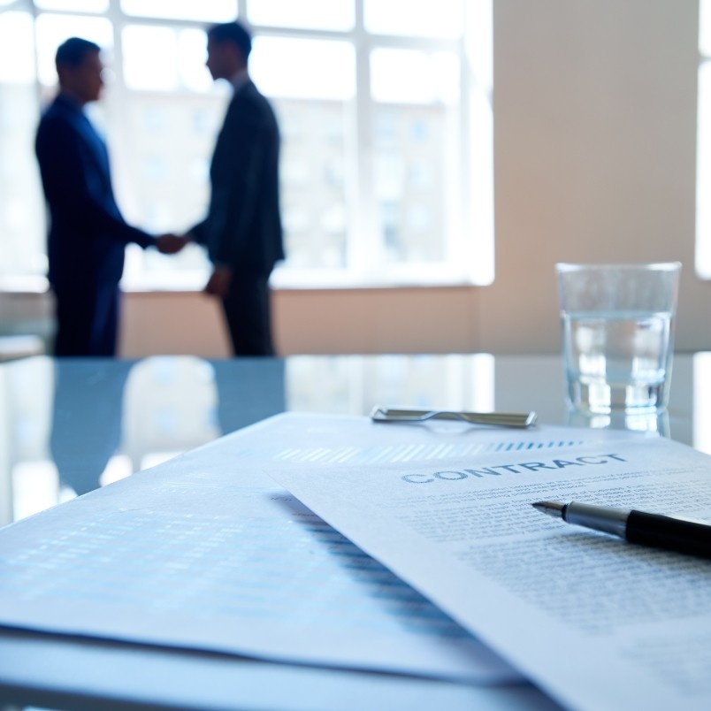 A contract and pen on a glass table in focus, symbolizing contract management, with two people in business attire shaking hands in the blurred background near large windows. A glass of water sits on the table.