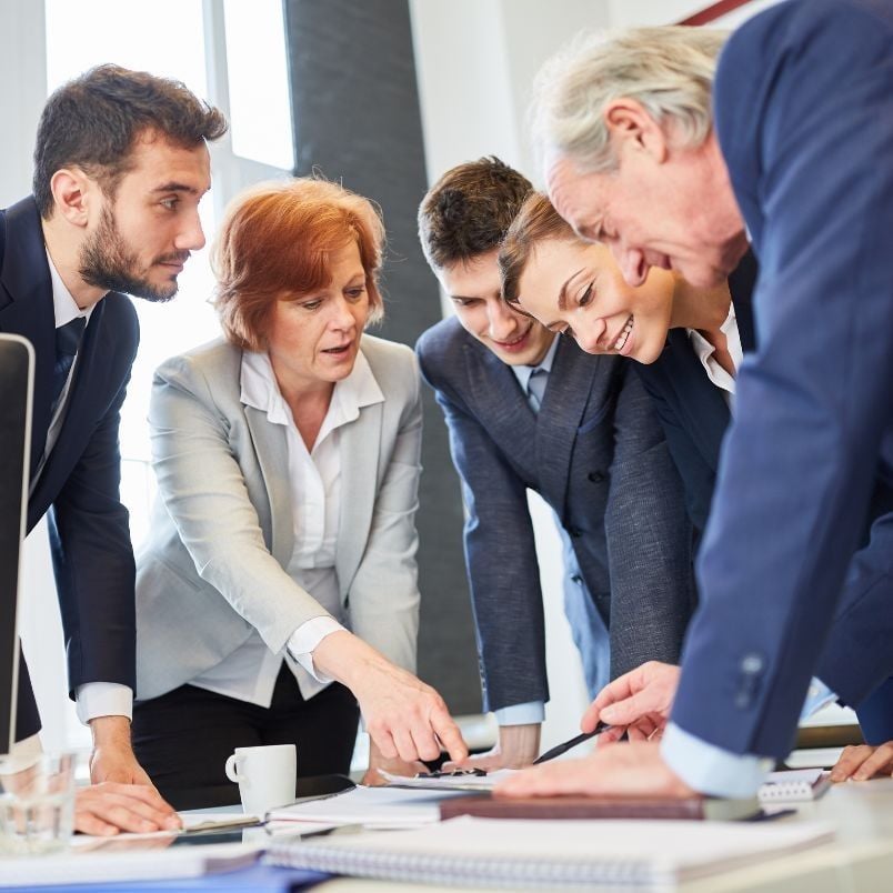 Five business professionals in formal attire are gathered around a table, focused on documents—such as an Agile Project Charter Template—and discussing work together in a bright office setting.