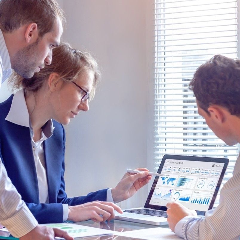 Three business professionals review charts on paper and analytical graphs on a laptop in a bright office, collaborating and discussing data results and a Wireframe Template by the window.