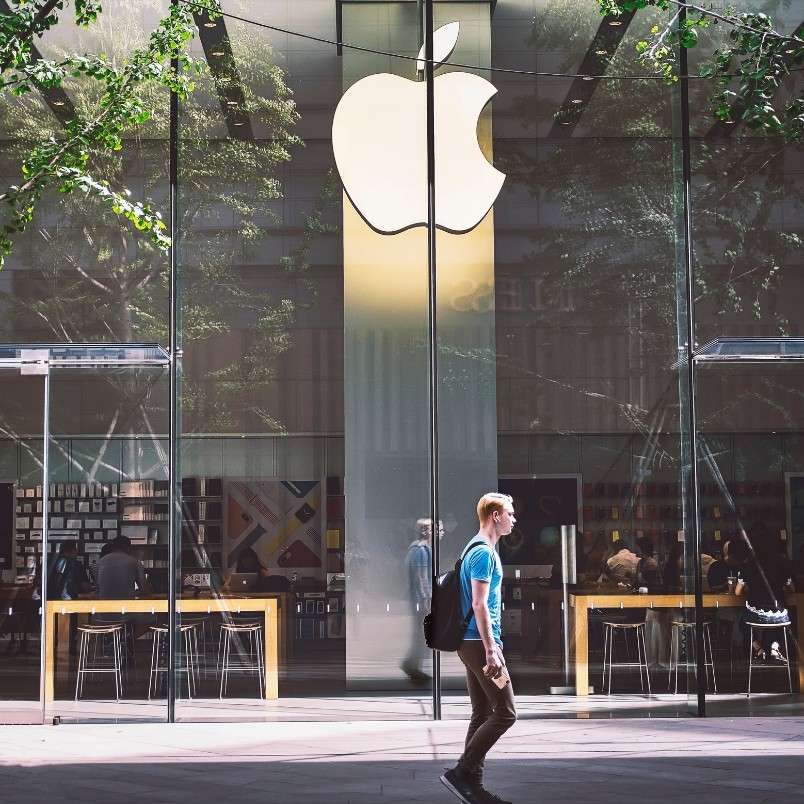 A person with a backpack walks past a glass-fronted Apple Store, its large white logo above the entrance—a scene that could serve as an agile project management example, with reflections and trees visible on the sleek glass exterior.
