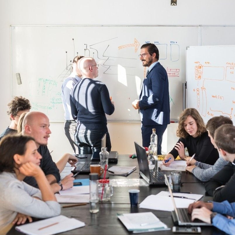 A group of people in a bright office collaborate around a table with laptops and papers, while three individuals stand at a whiteboard discussing diagrams and notes for a new features template.
