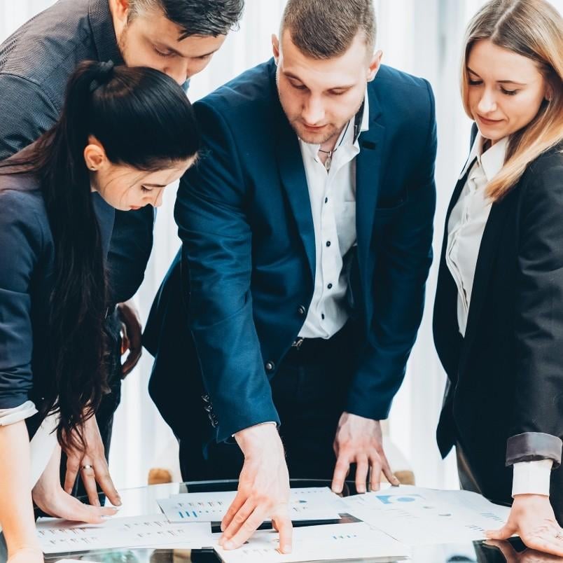 Four business professionals gather around a table, closely examining charts and graphs as they discuss the project manager role in Scrum within a bright, collaborative office setting.