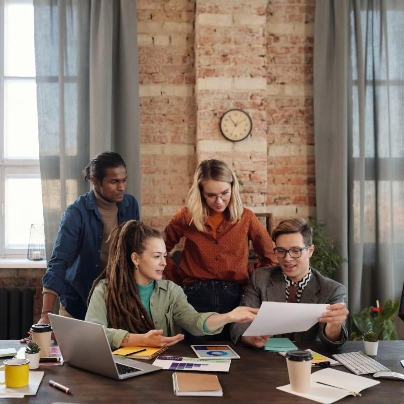Four people gather around a desk with laptops and papers, actively discussing an Agile Team Charter. Two are seated while two stand behind them. The background features brick walls, tall curtains, and a clock.