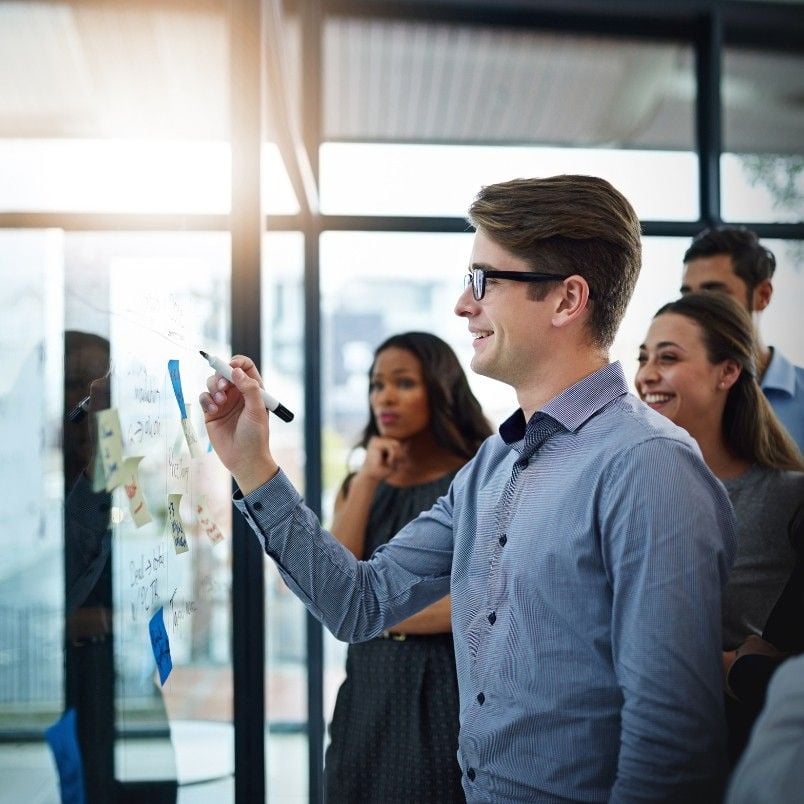 A man in glasses writes on a glass board with a marker, outlining the project charter while a group of colleagues stand nearby smiling and watching in a modern agile office setting.
