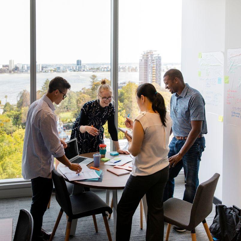 Four people stand around a round table in a modern office with large windows, collaborating on documents, a laptop, and an Agile Project Charter Template. A cityscape and water are visible outside. Charts and notes are on the wall.