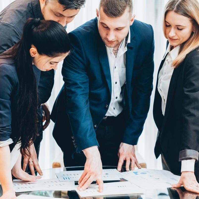 Four business professionals in formal attire are gathered around a table, closely reviewing documents and charts that showcase a features template. One person is pointing to a paper as the group engages in focused discussion and analysis.