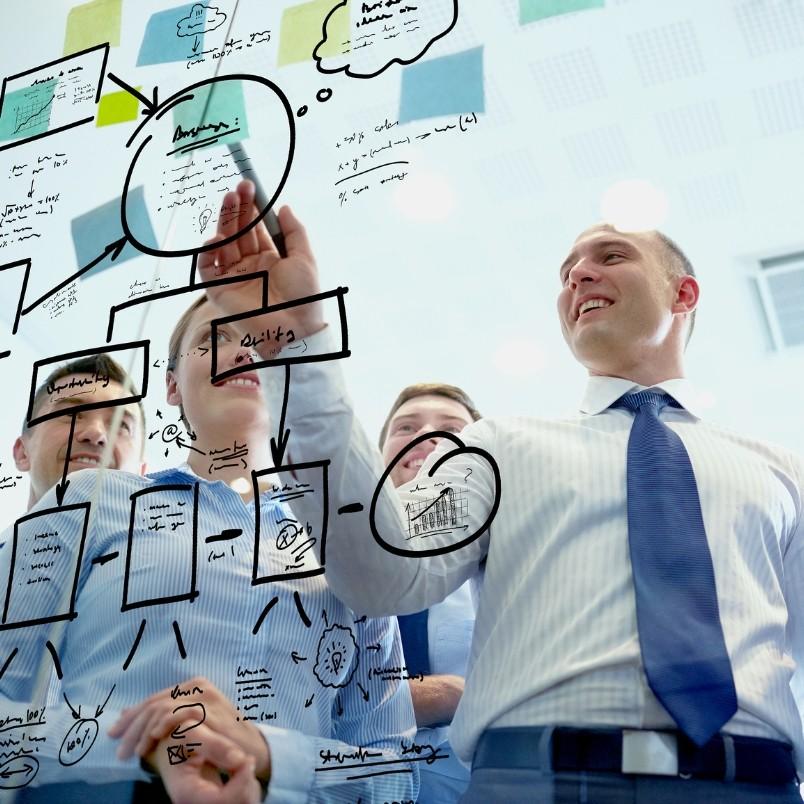 Four people in business attire smile and collaborate, brainstorming ideas as they write diagrams, graphs, and notes with black marker on a glass wall in a modern office setting.