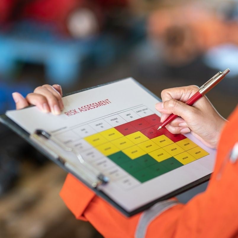 A person in an orange sleeve holds a clipboard with a “Risk Assessment” chart and risk register, using a red pen to mark the colored grid of green, yellow, and red zones. The background is blurred.